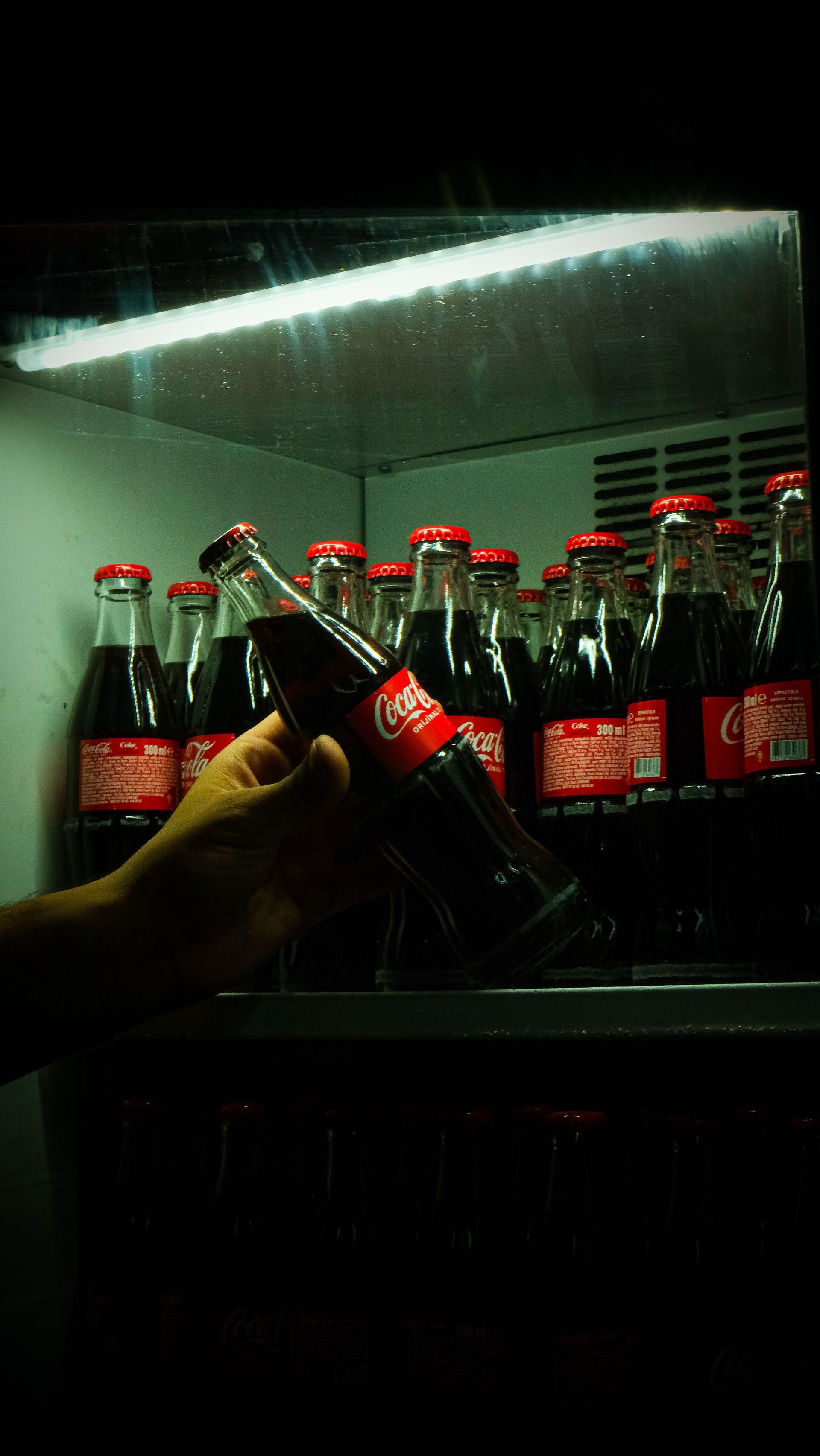Hand holding a Coca-Cola bottle in front of a cooler filled with red-capped soda bottles