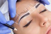Person's face with eyebrows marked for shaping. A gloved hand holds a pencil, skin is visible.