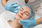Woman receiving a facial injection; gloved hands, blue gloves, white cotton pad, and a medical setting.