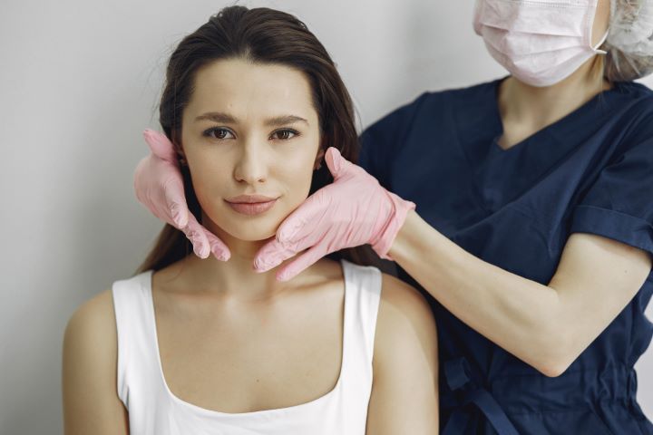 Woman having her face examined by a person wearing medical attire and gloves.