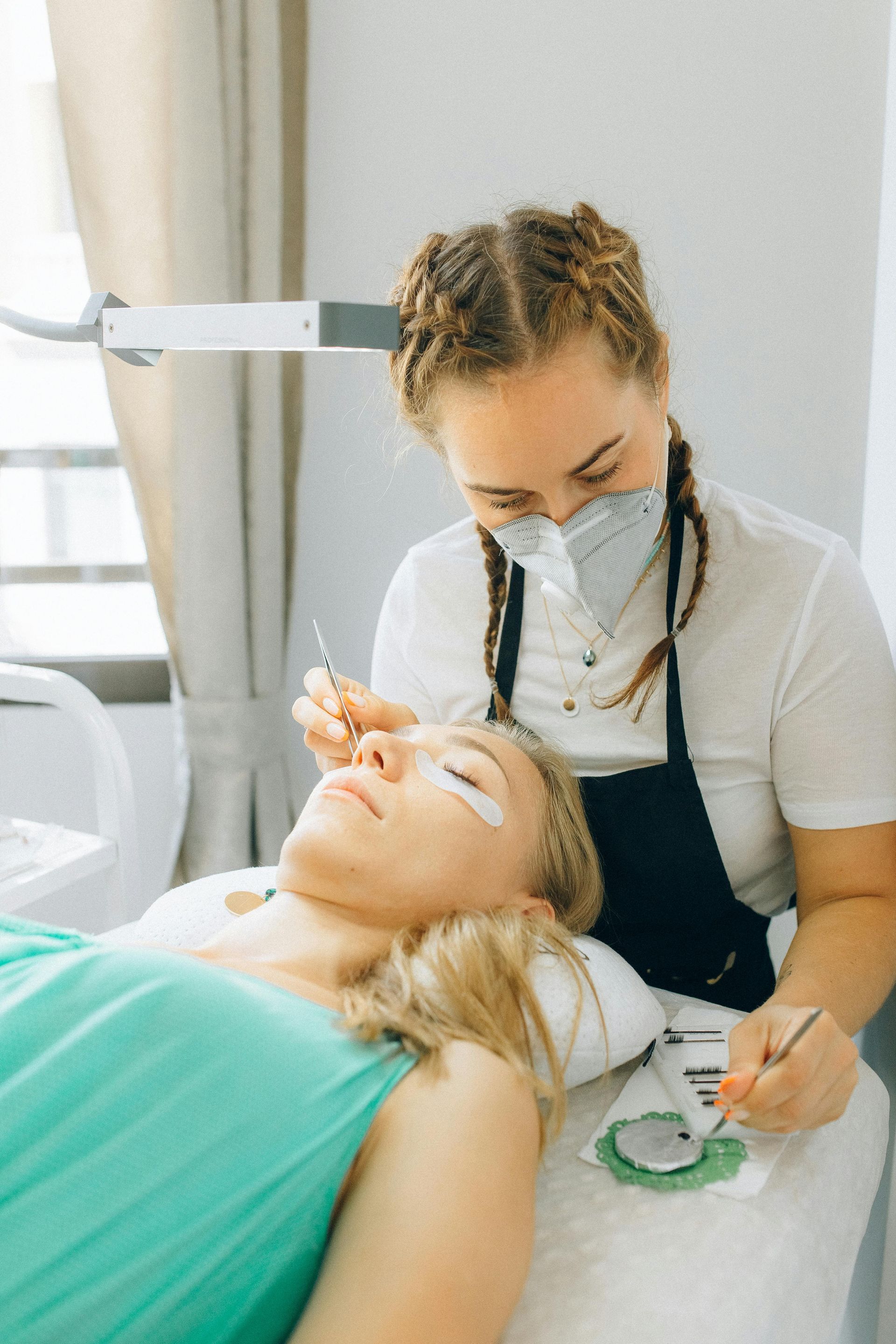 Woman getting eyelash extensions applied by a technician wearing a mask, white room.
