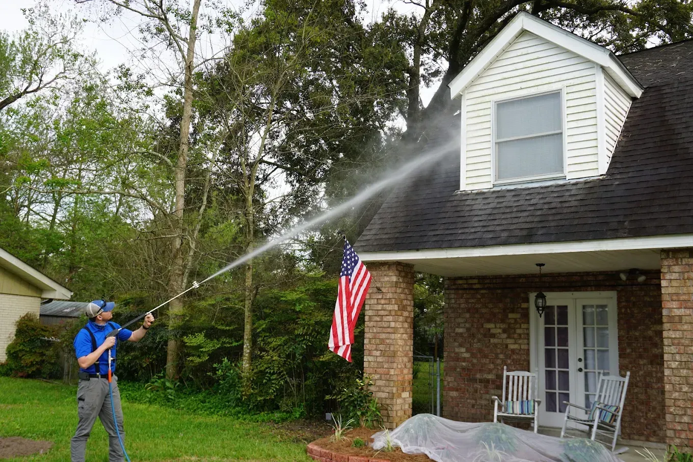 Man spraying water from a hose at a brick house with an American flag hanging by the porch
