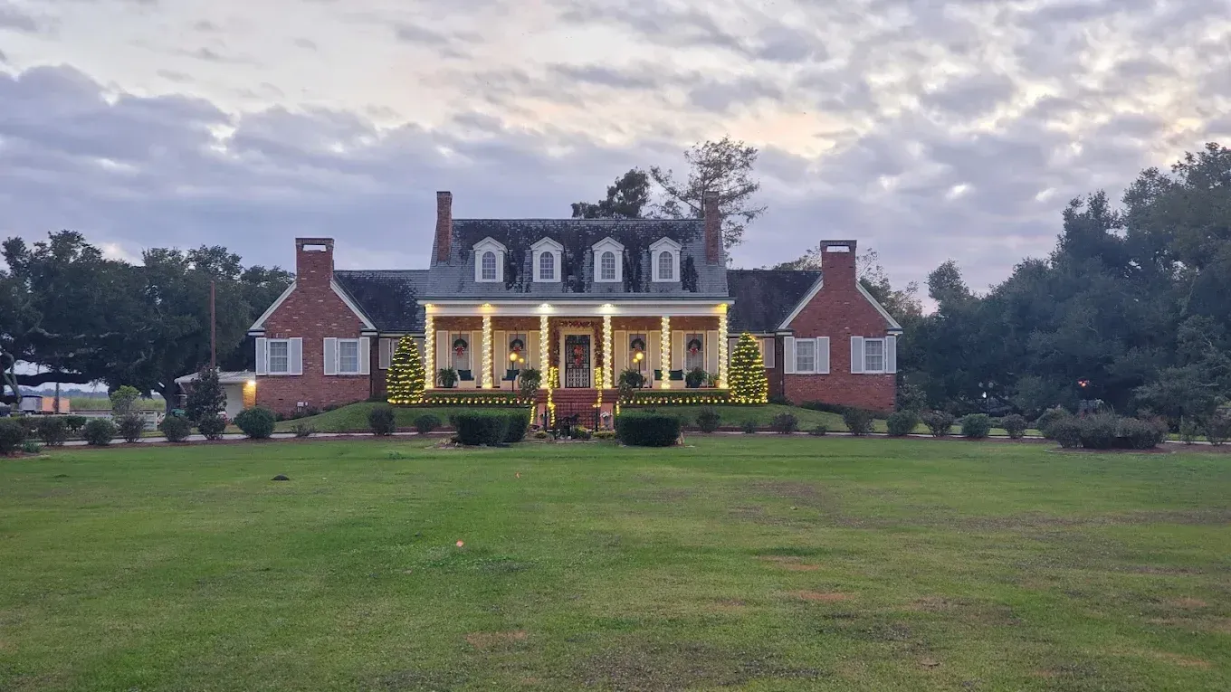 Large colonial-style house with lit porch and red brick wings, seen across a wide green lawn at dusk