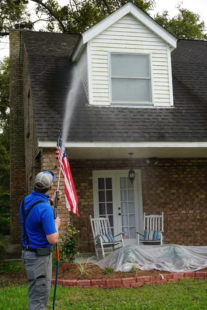 Person spraying water on a house roof beside an American flag and front porch.