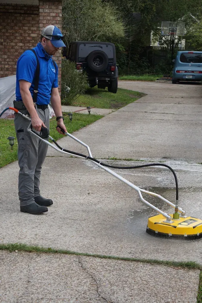 Worker pressure-washing a concrete driveway with a yellow surface cleaner attachment