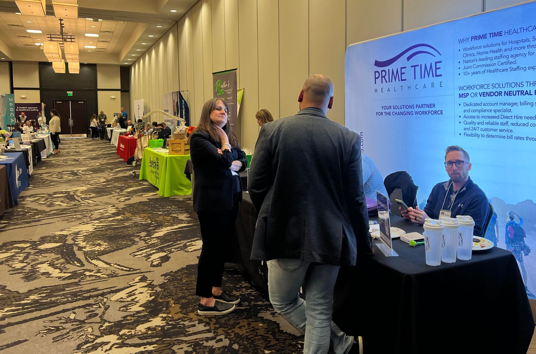 Conference hall with people at vendor booths, including a person speaking with a vendor at a