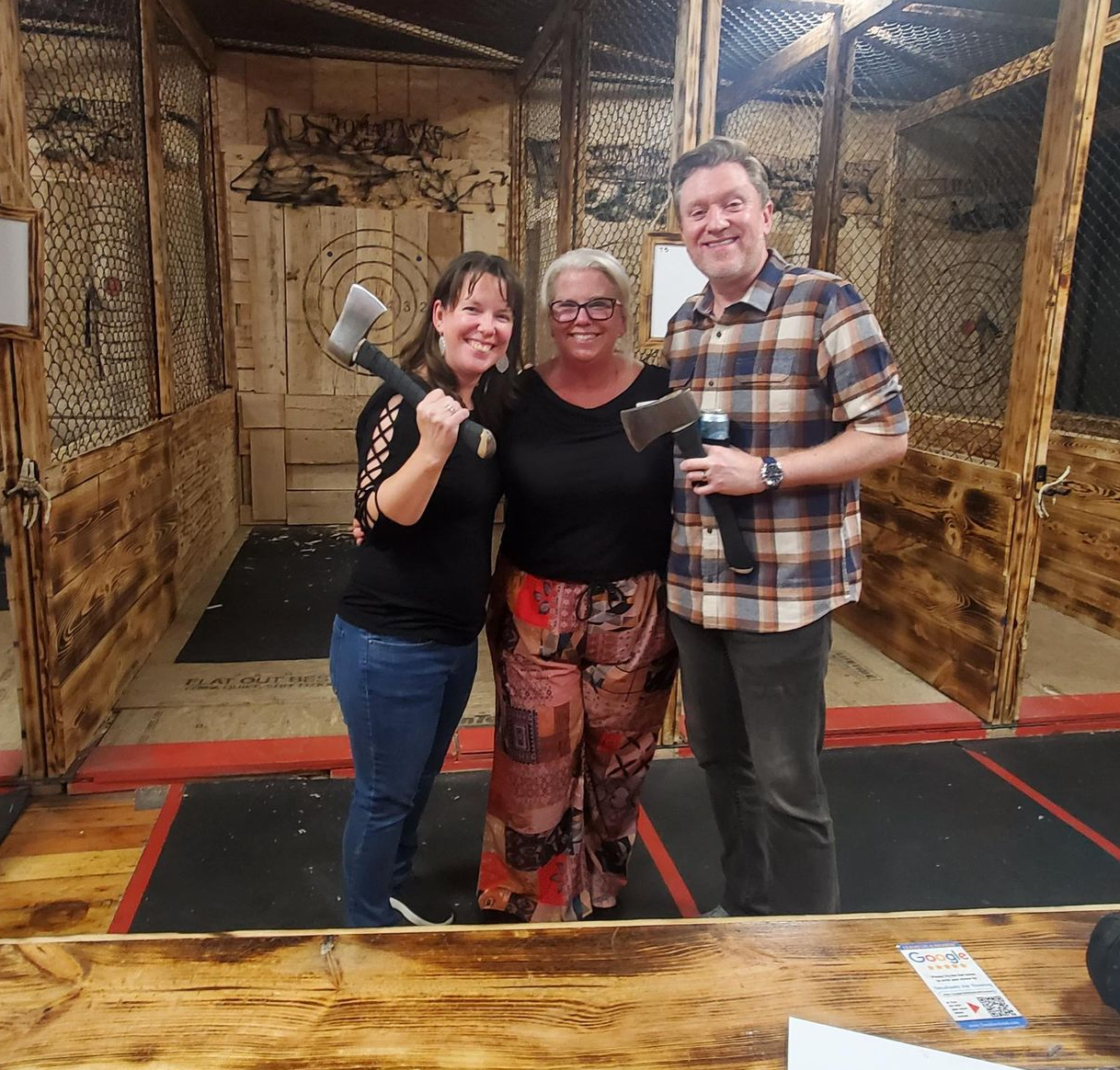 Three people pose at an axe-throwing range: Two women holding axes smile; a man holds a microphone and smiles.
