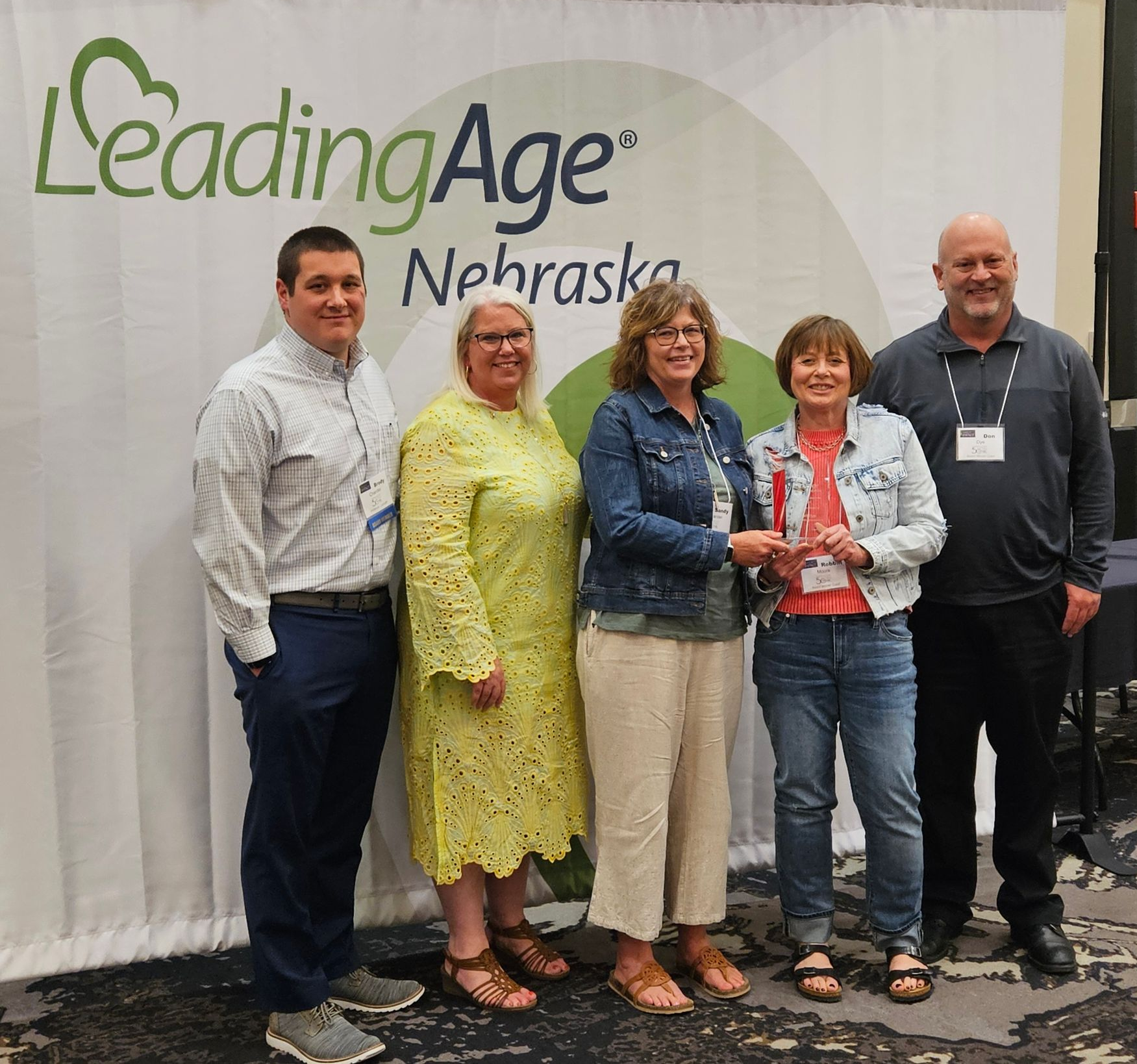 Five people posing in front of a LeadingAge Nebraska banner, two women holding a trophy.