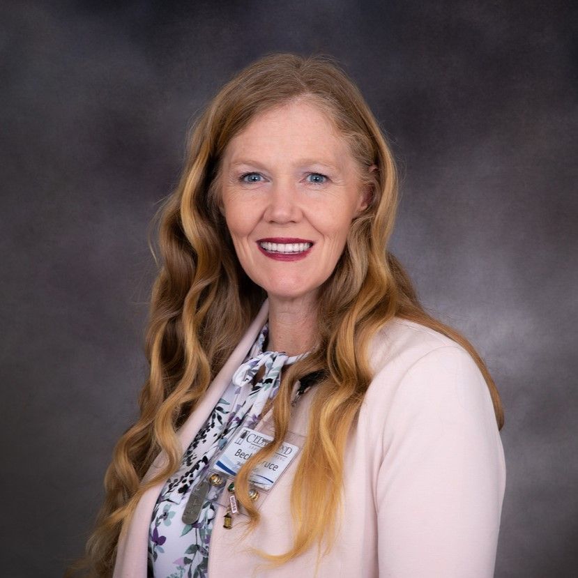 Woman with long wavy blonde hair, wearing a pink blazer over a floral top, smiling at the camera.