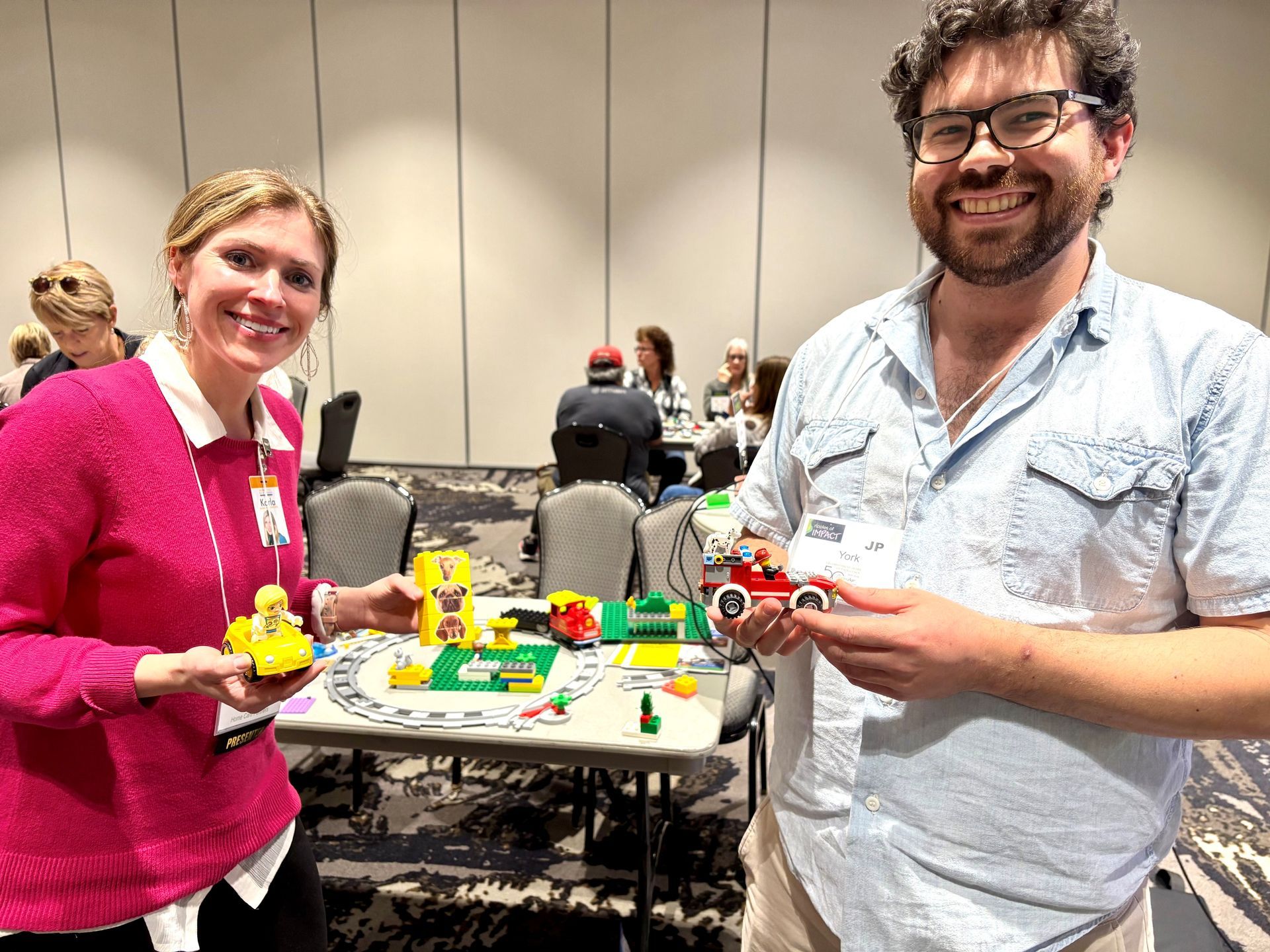 Two people smiling, holding Lego creations, in a conference room with others.