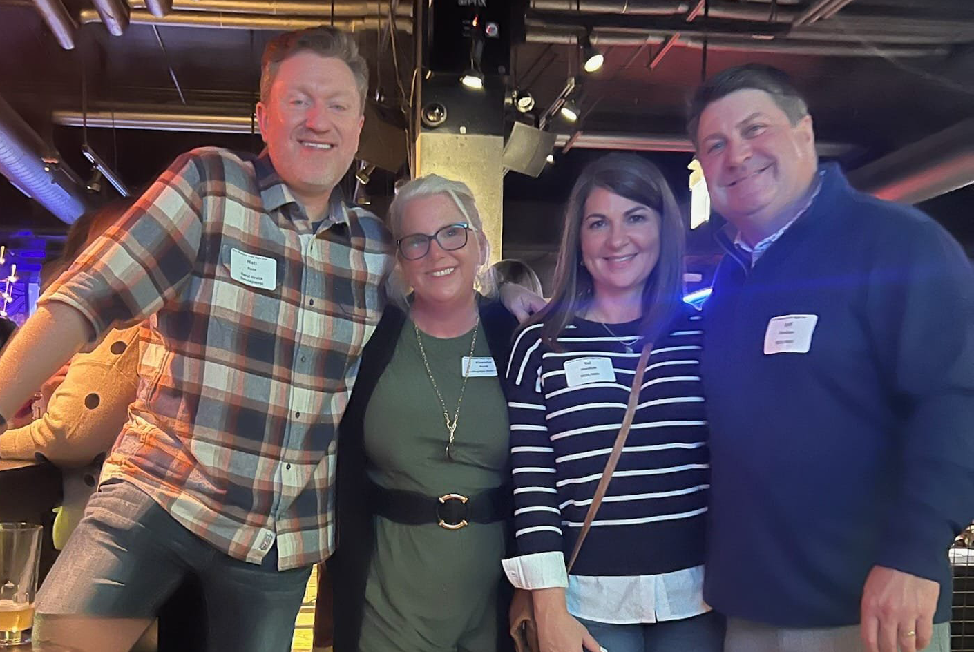Four people smiling and posing together indoors. A man in a plaid shirt and another in a blue sweater stand on either side of two women.