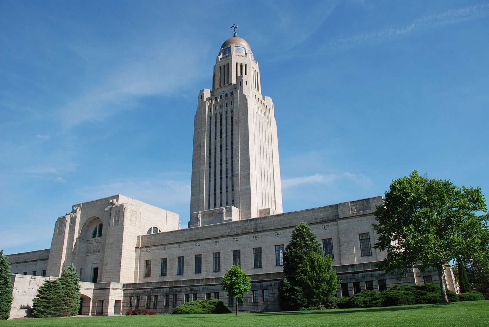 Nebraska State Capitol Building against a blue sky. Tall tower with clock, light-colored stone.