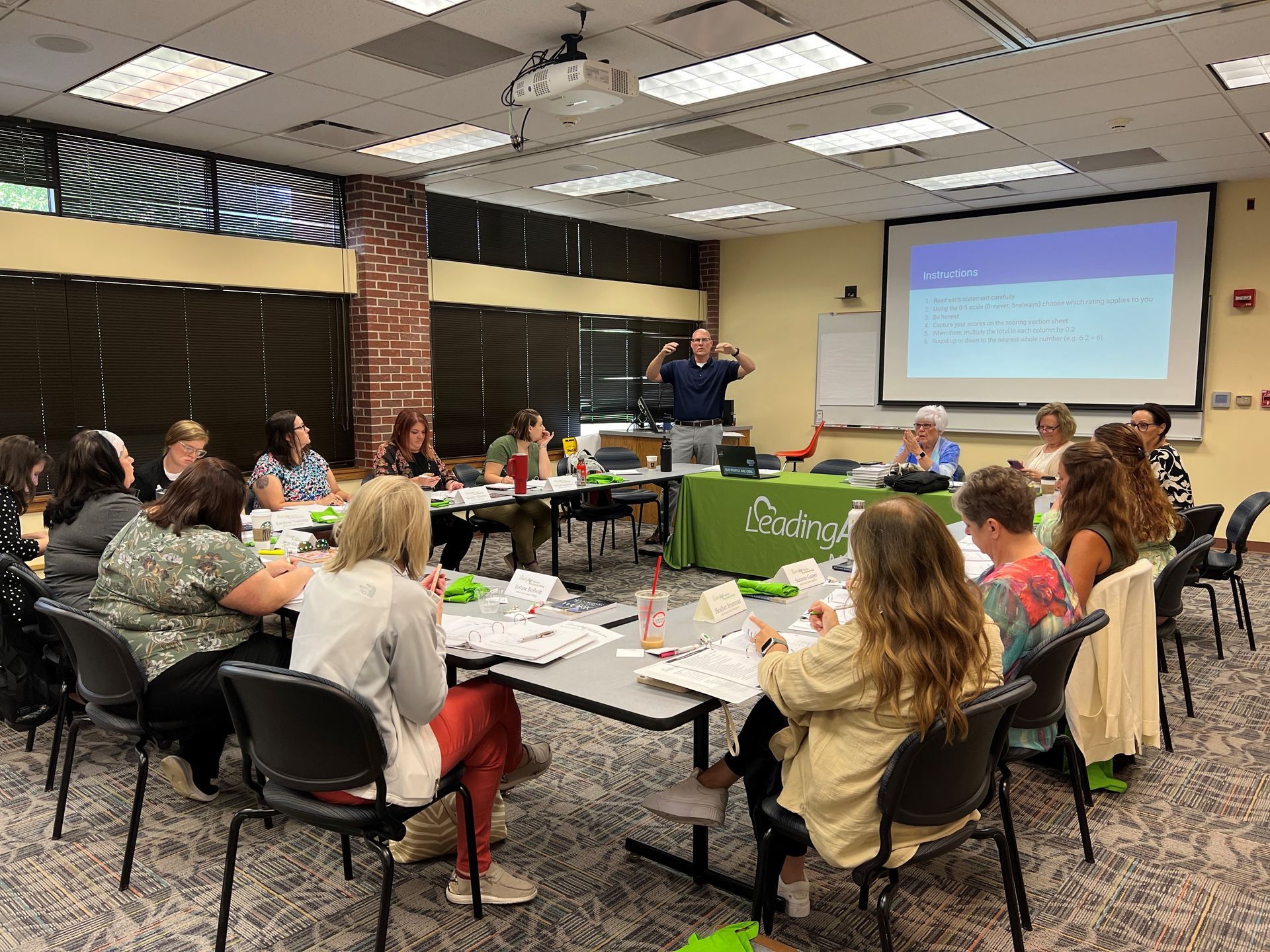 A group of people sitting around tables, attending a training session; presenter at the front.