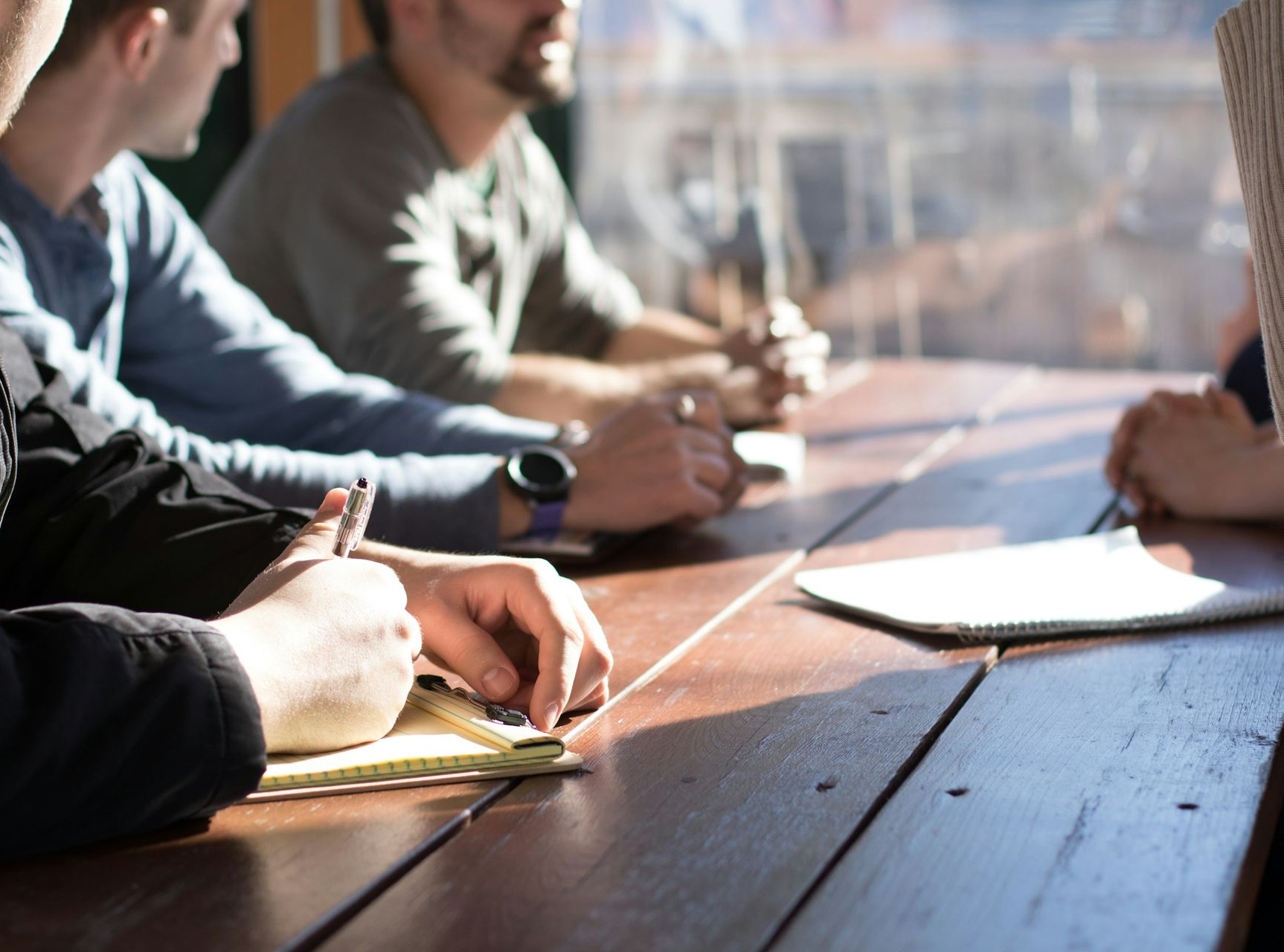 People at a table writing in notebooks. Sunlight streams across the wooden surface.