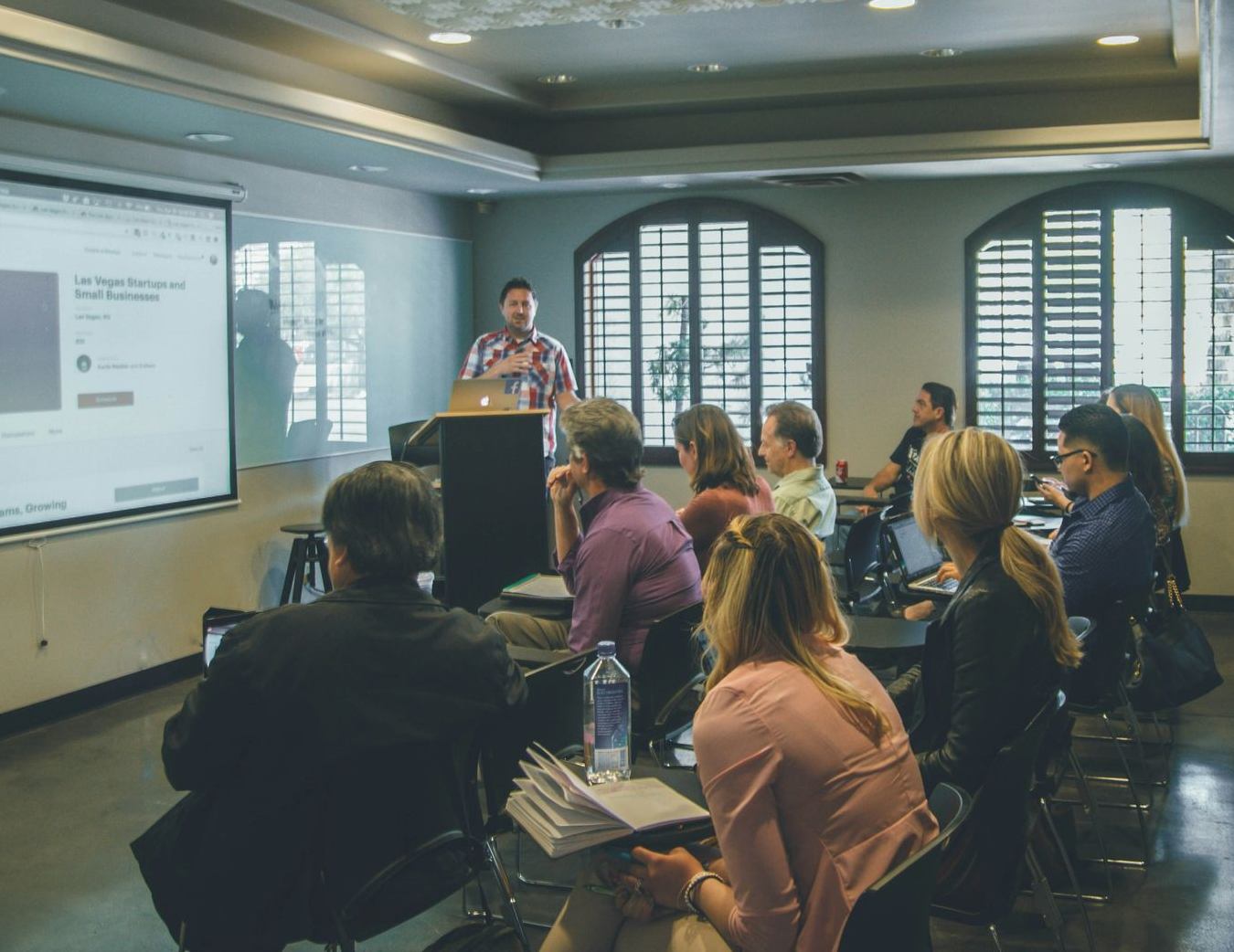 Man lecturing to a group in a room. Projector screen visible, audience seated in chairs.