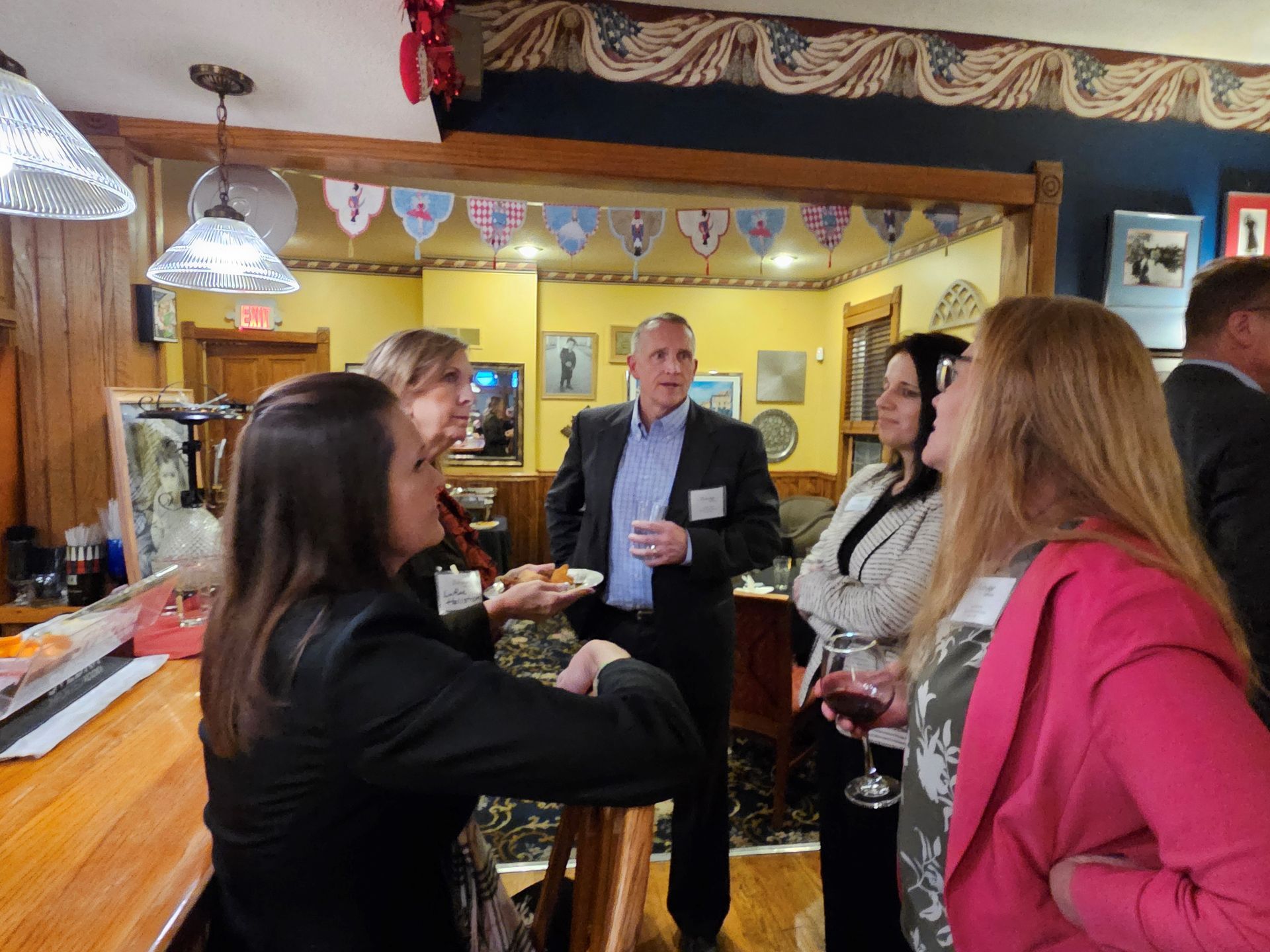 Group of people at a gathering in a room with a bar, some holding drinks and food, talking.