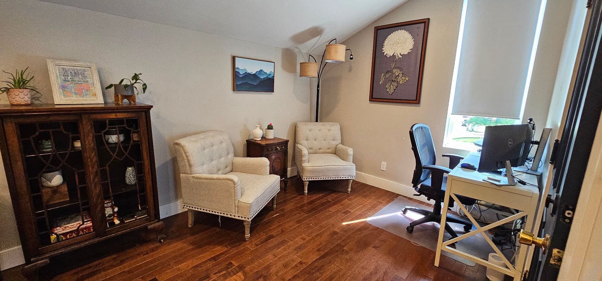 A room with two armchairs, a desk, a cabinet, and a framed botanical print. Sunlight streams in from a window.