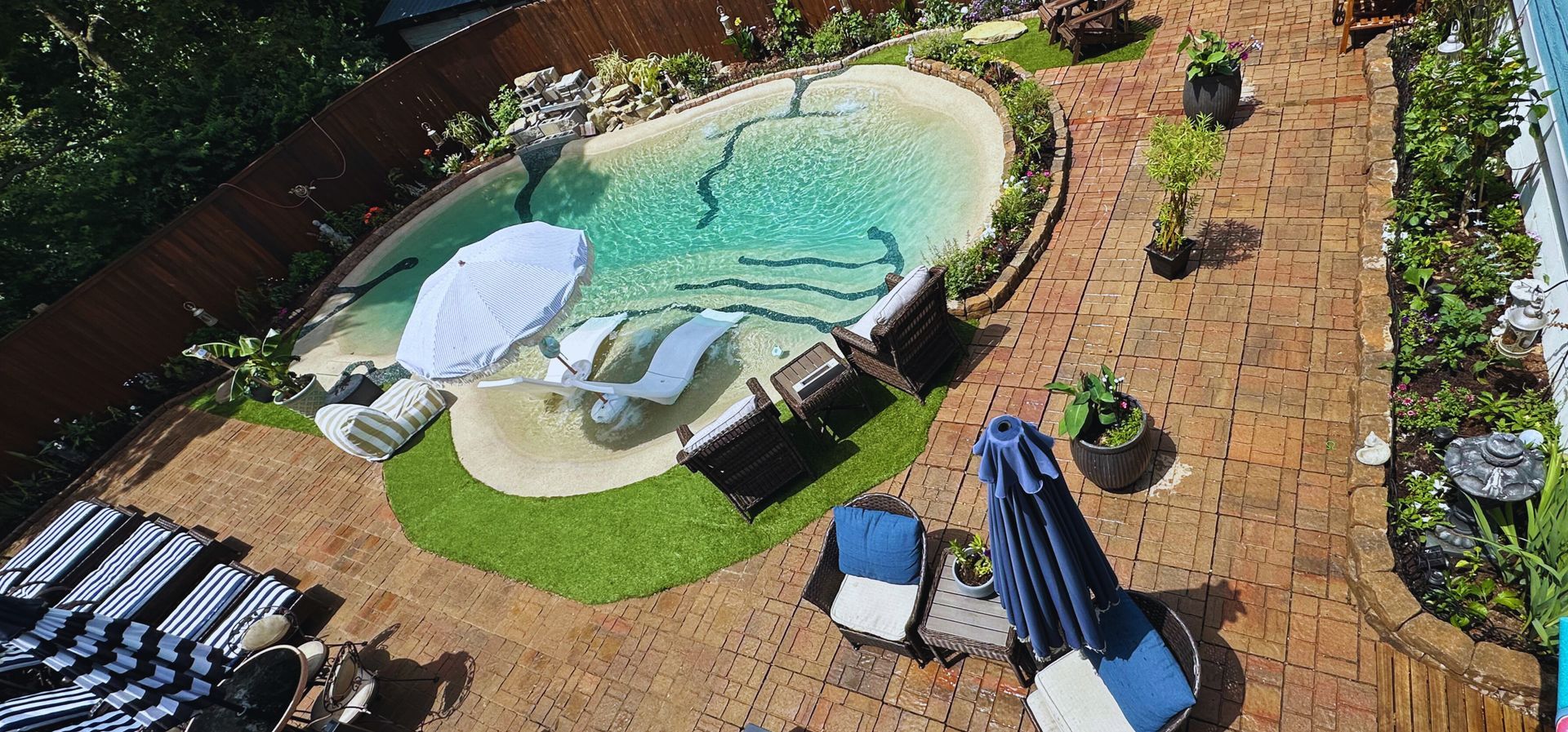 Aerial view of a backyard pool surrounded by brick patio, lawn, and greenery with lounge chairs and umbrellas.