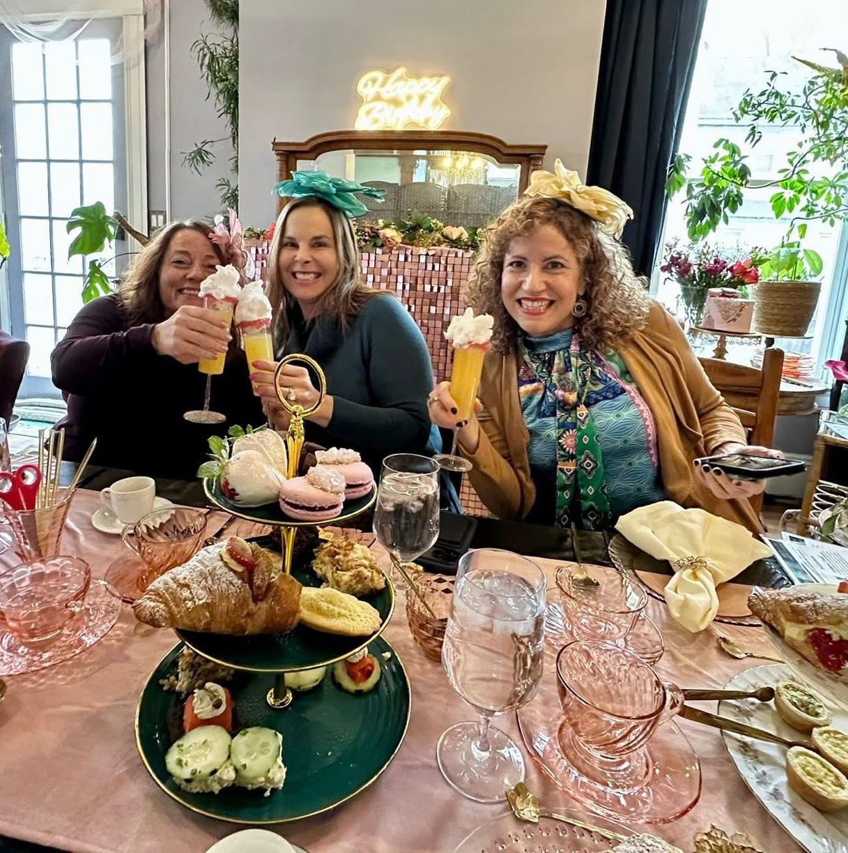 Three friends toasting drinks at a café table with pastries and flowers in a bright dining room