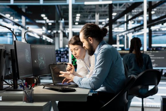 Two coworkers reviewing a tablet at desks in an open office with computer monitors and glass walls.