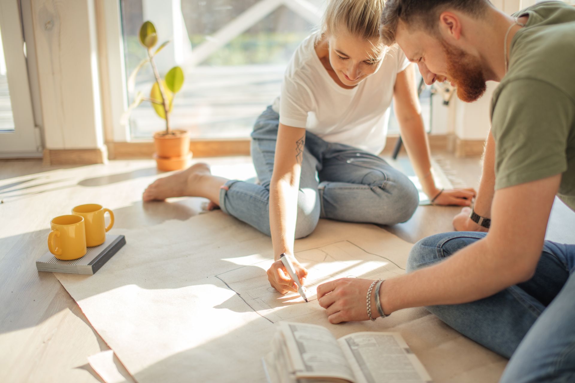 Couple in casual clothes, sketching on paper on the floor near window, considering home design.