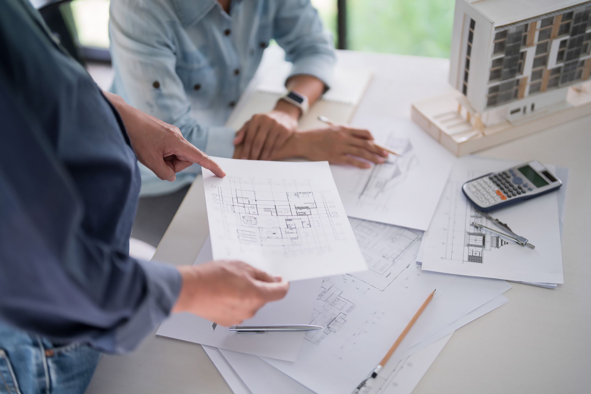 Two people reviewing architectural blueprints at a desk with a model building and calculator.