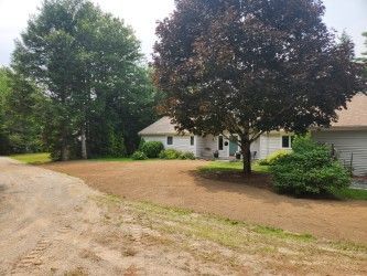 Driveway beside a white house with large trees and a gravel road in front
