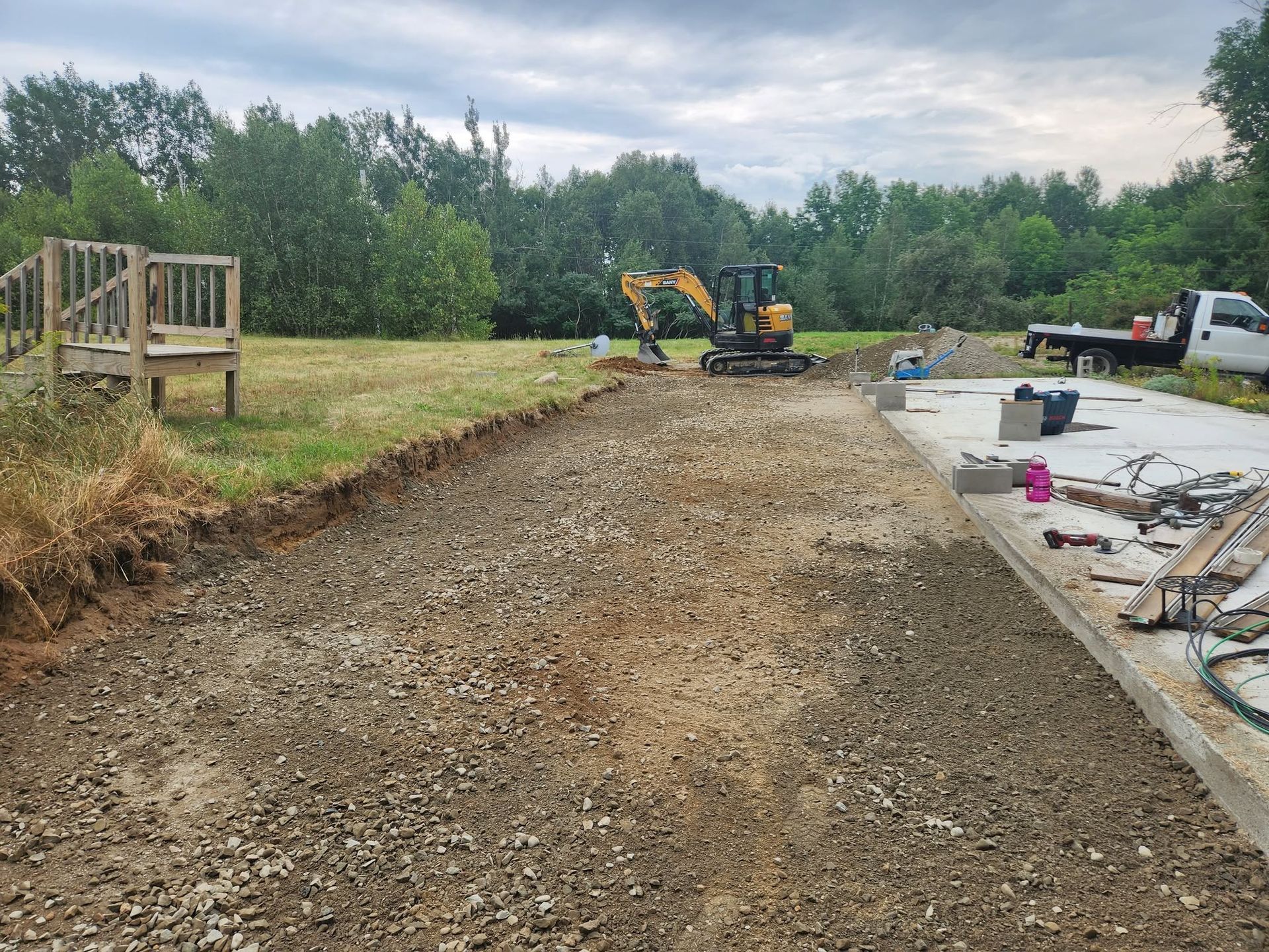 Construction site with gravel driveway, excavator, and workers’ materials beside trees