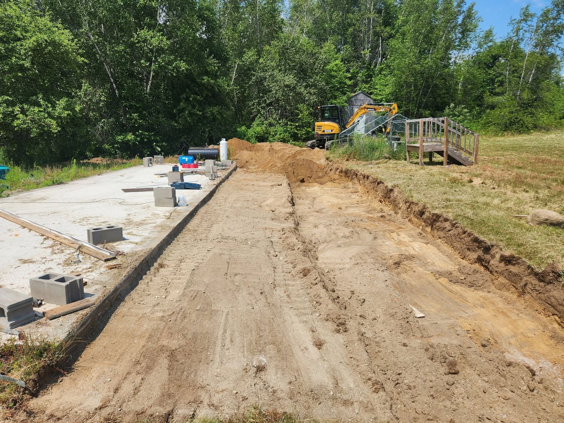 Partially built dirt path beside a concrete walkway with construction equipment and trees in the background