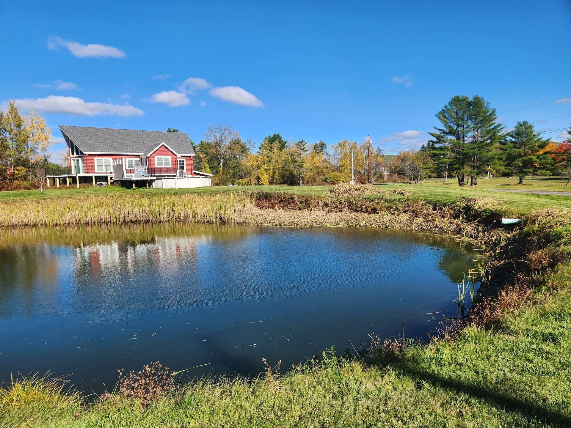 Red house by a pond under a blue sky, with autumn trees and grassy banks