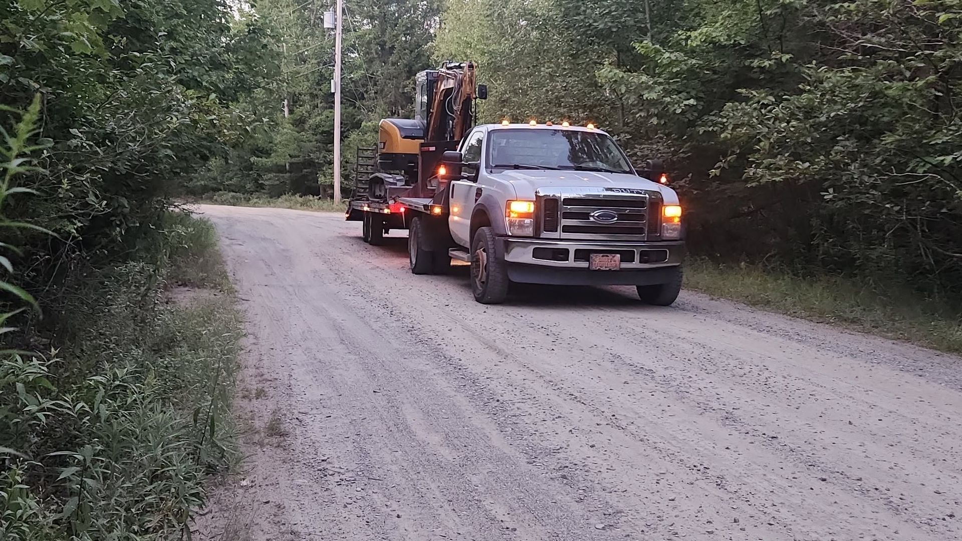 Pickup truck hauling construction equipment on a gravel road in a wooded area