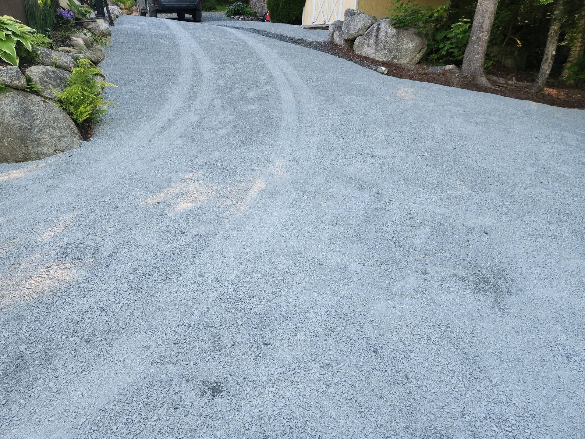 Gravel driveway curving uphill between rocks and trees, with a vehicle in the distance