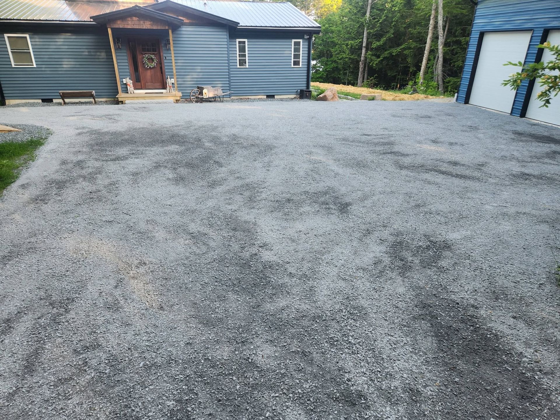 Gravel driveway leading to a blue house with a front porch and garage, surrounded by trees