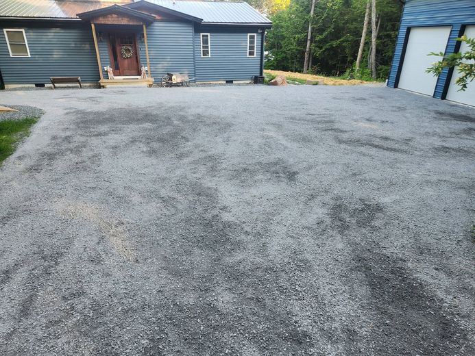 Gravel driveway leading to a blue house with a front porch and garage, surrounded by trees