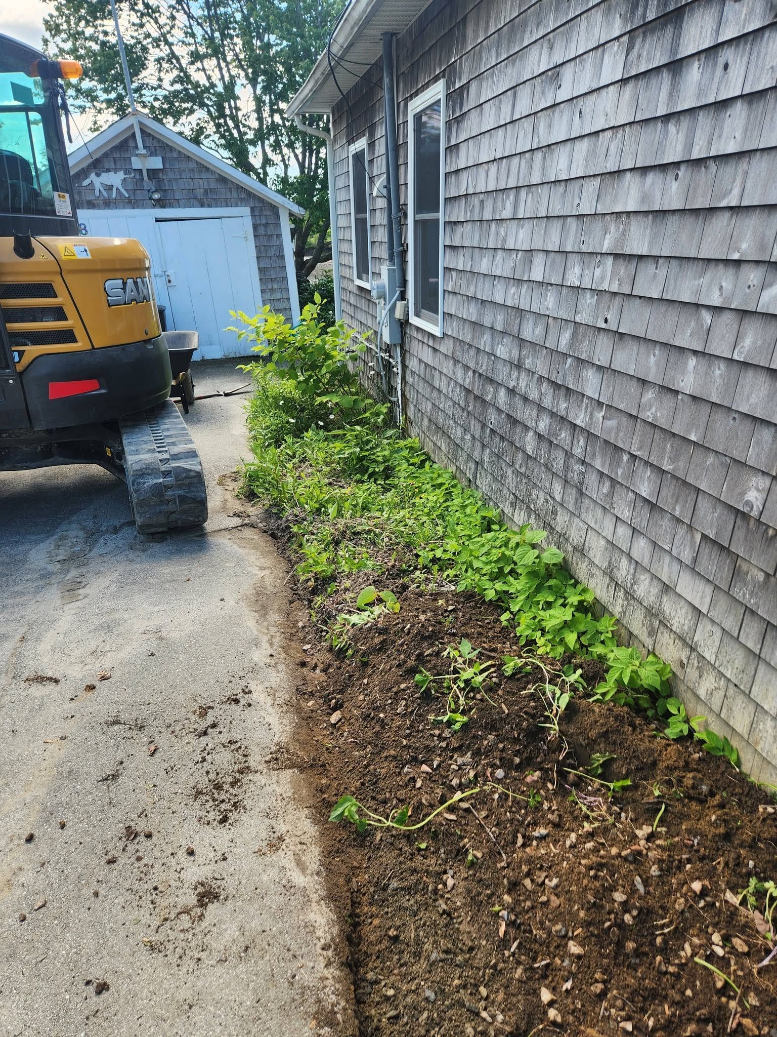 Side yard beside a gray shingle house, with mulch beds and a yellow utility truck parked on the driveway.