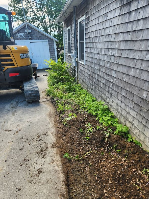 Narrow driveway beside shingled house, with a yellow construction vehicle and a mulched garden bed along the wall