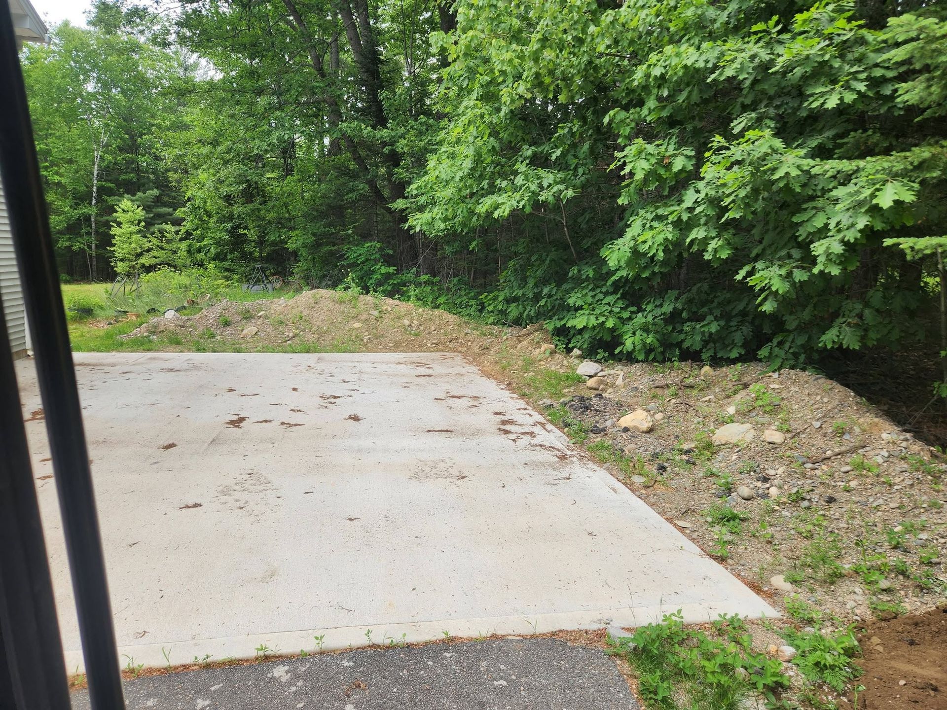 Concrete slab beside a wooded dirt slope with green trees and shrubs