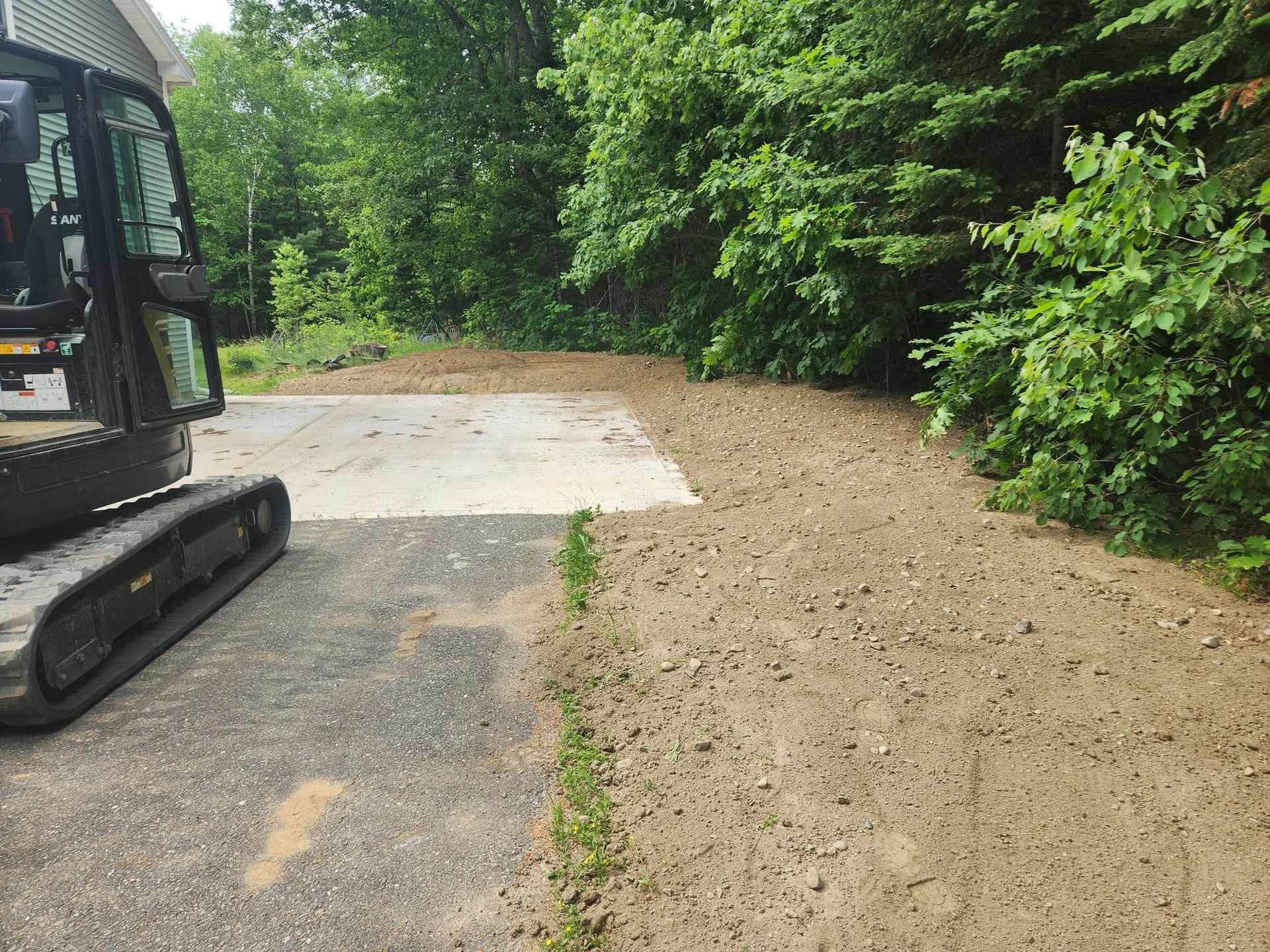 Excavator beside a freshly cleared dirt area next to pavement and dense green trees