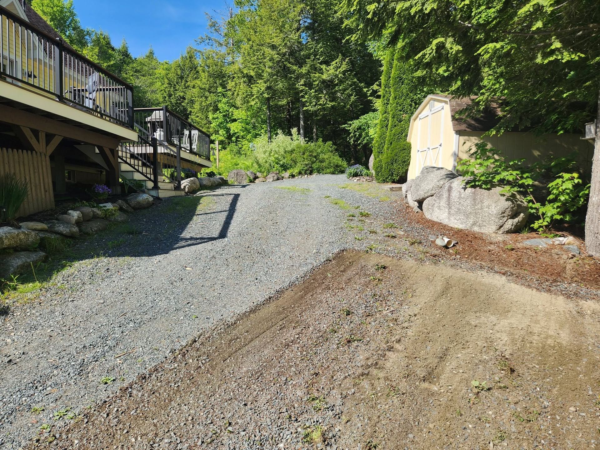 Gravel driveway beside houses and trees on a sunny day, with a shed and retaining wall.