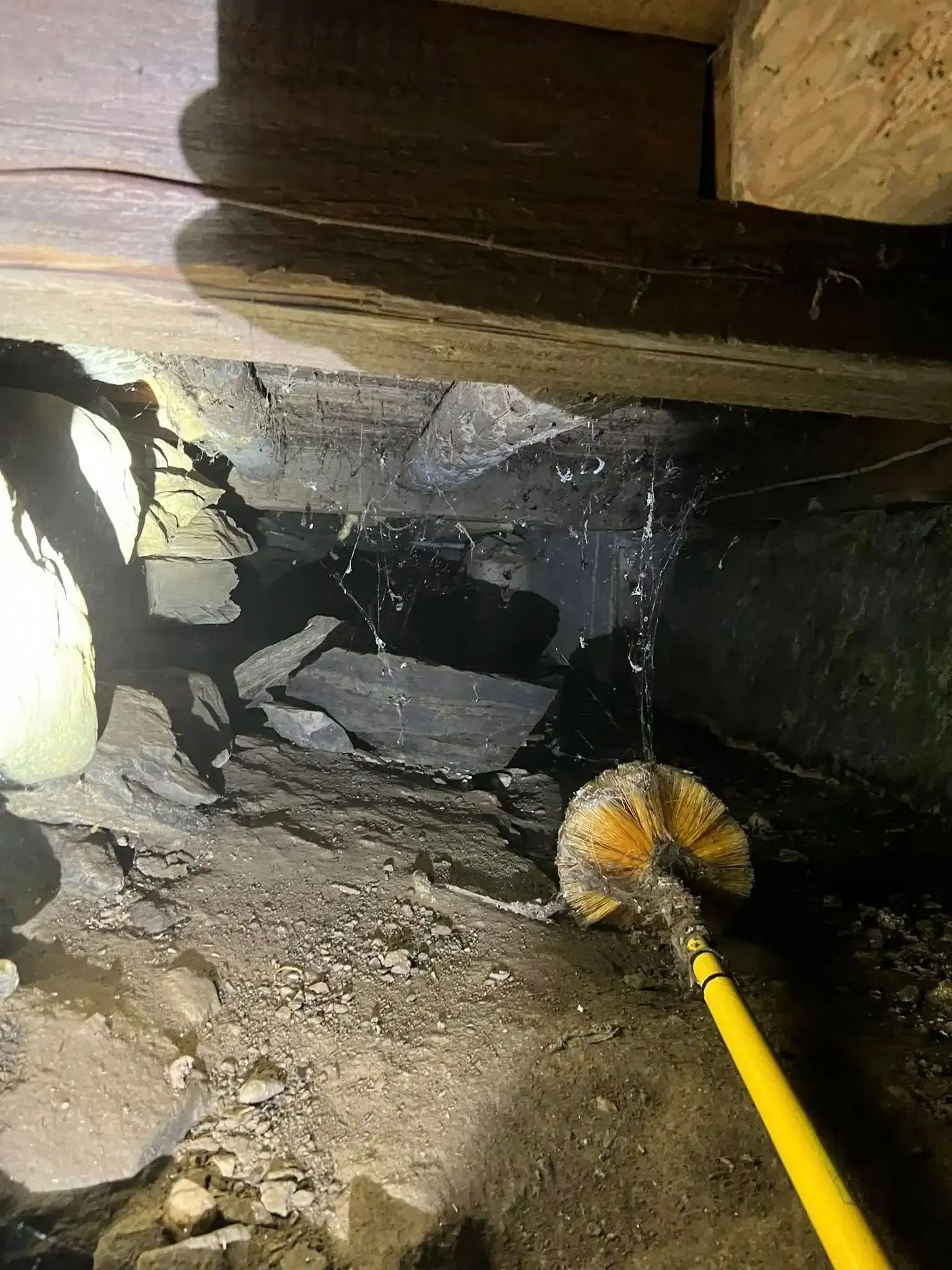 Yellow-handled brush sweeping debris under a dark wooden structure in a dusty crawlspace