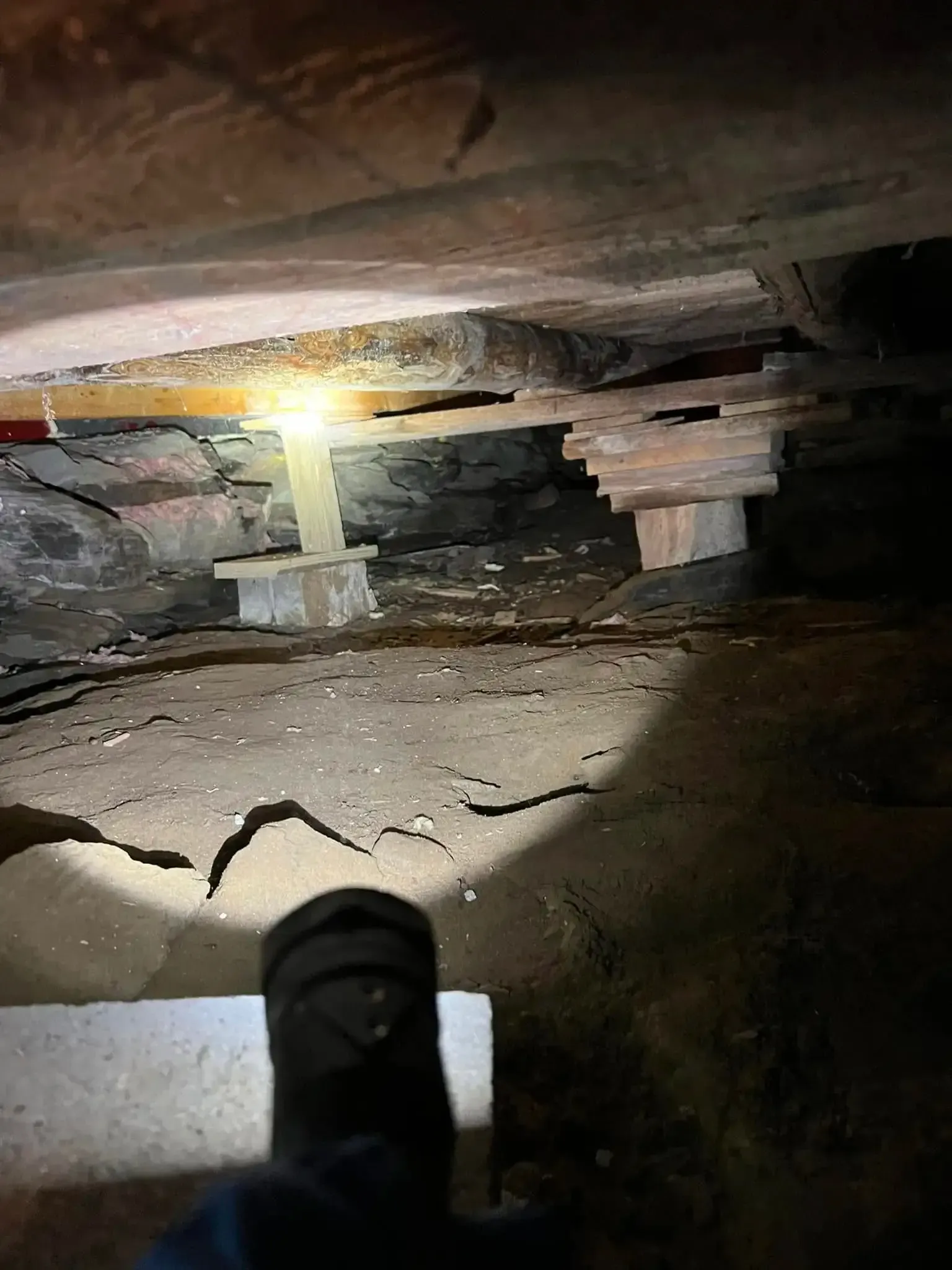 Flashlight illuminating a narrow crawlspace with rough beams and debris on the ground