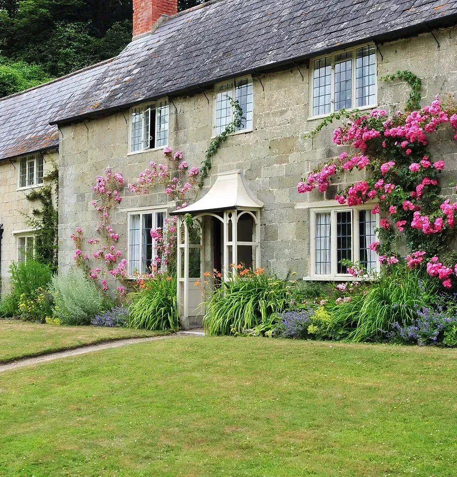 Stone cottage with pink climbing roses, white windows, and a green lawn in front