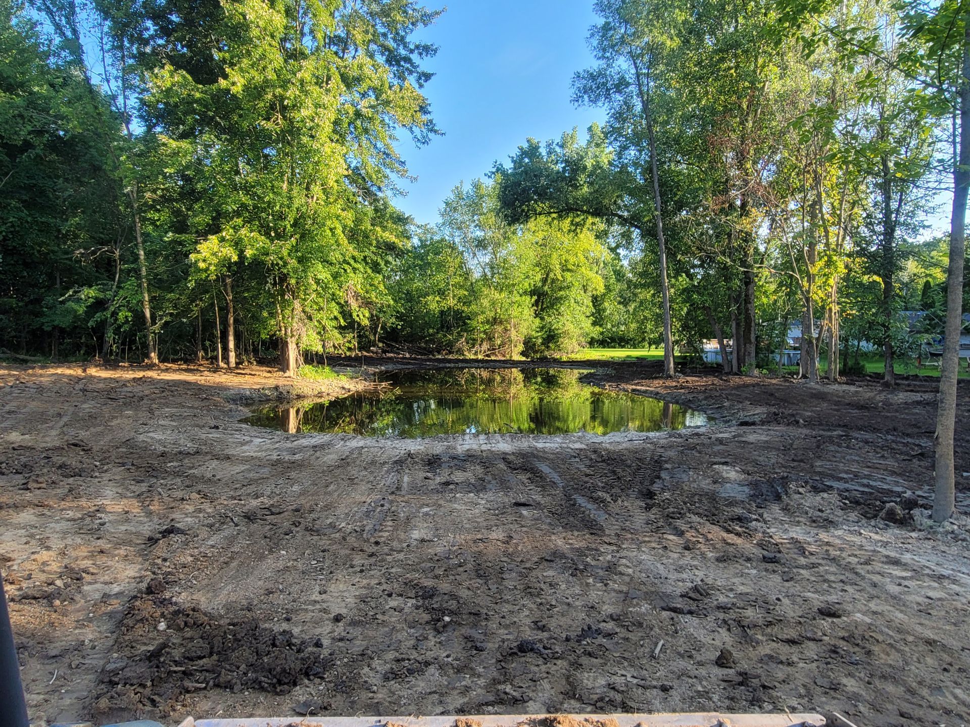 A cleared, muddy construction area surrounds a small pond nestled among trees under a bright blue sky.