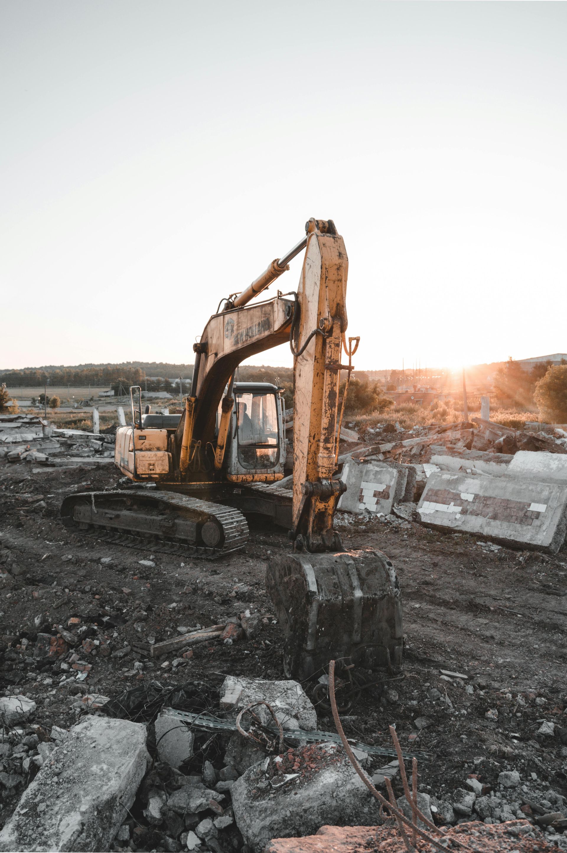 A yellow excavator sits in a rocky, debris-filled demolition site during a bright sunset.