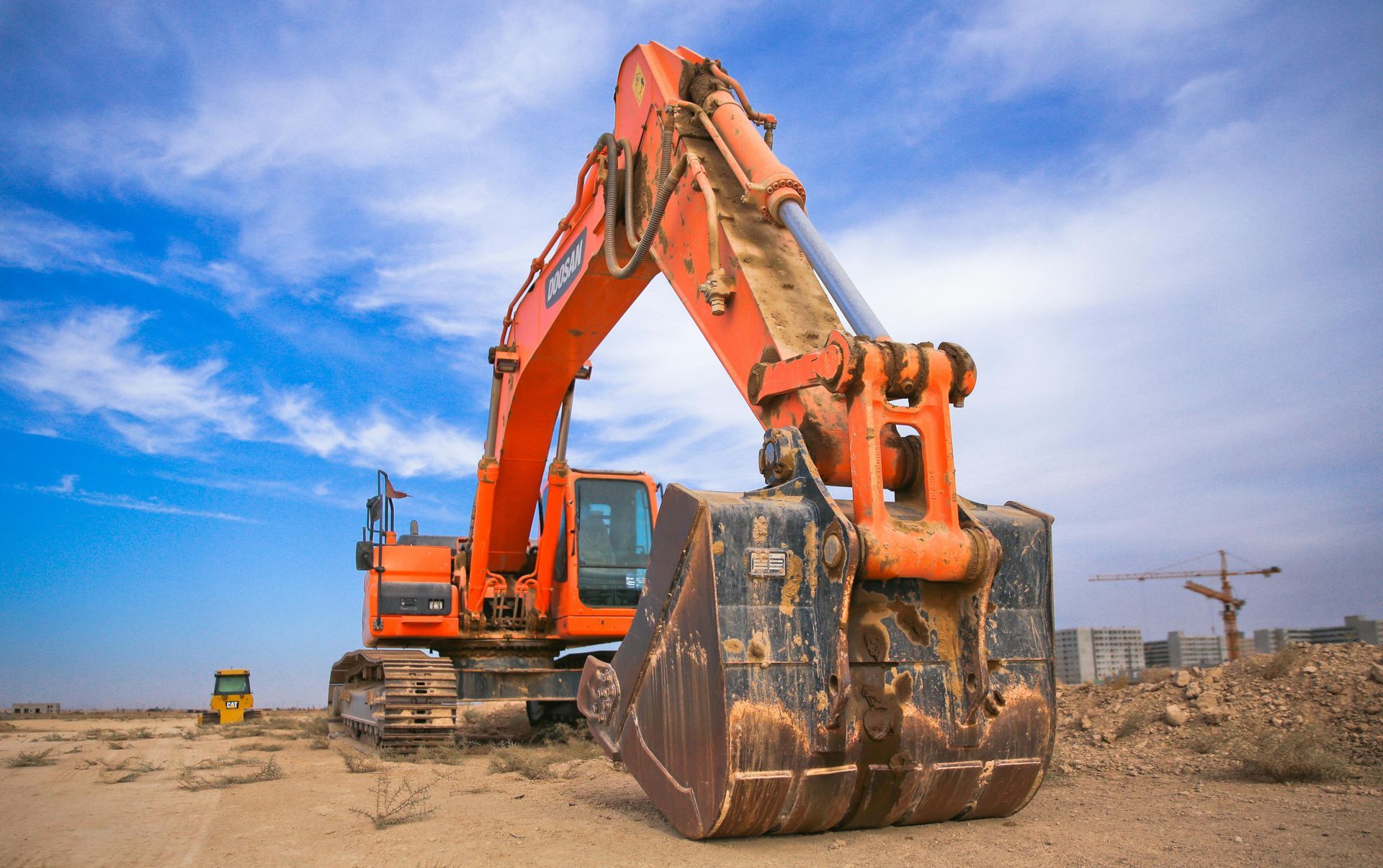 An orange excavator with a large metal bucket parked on a dirt construction site under a blue, cloudy sky.