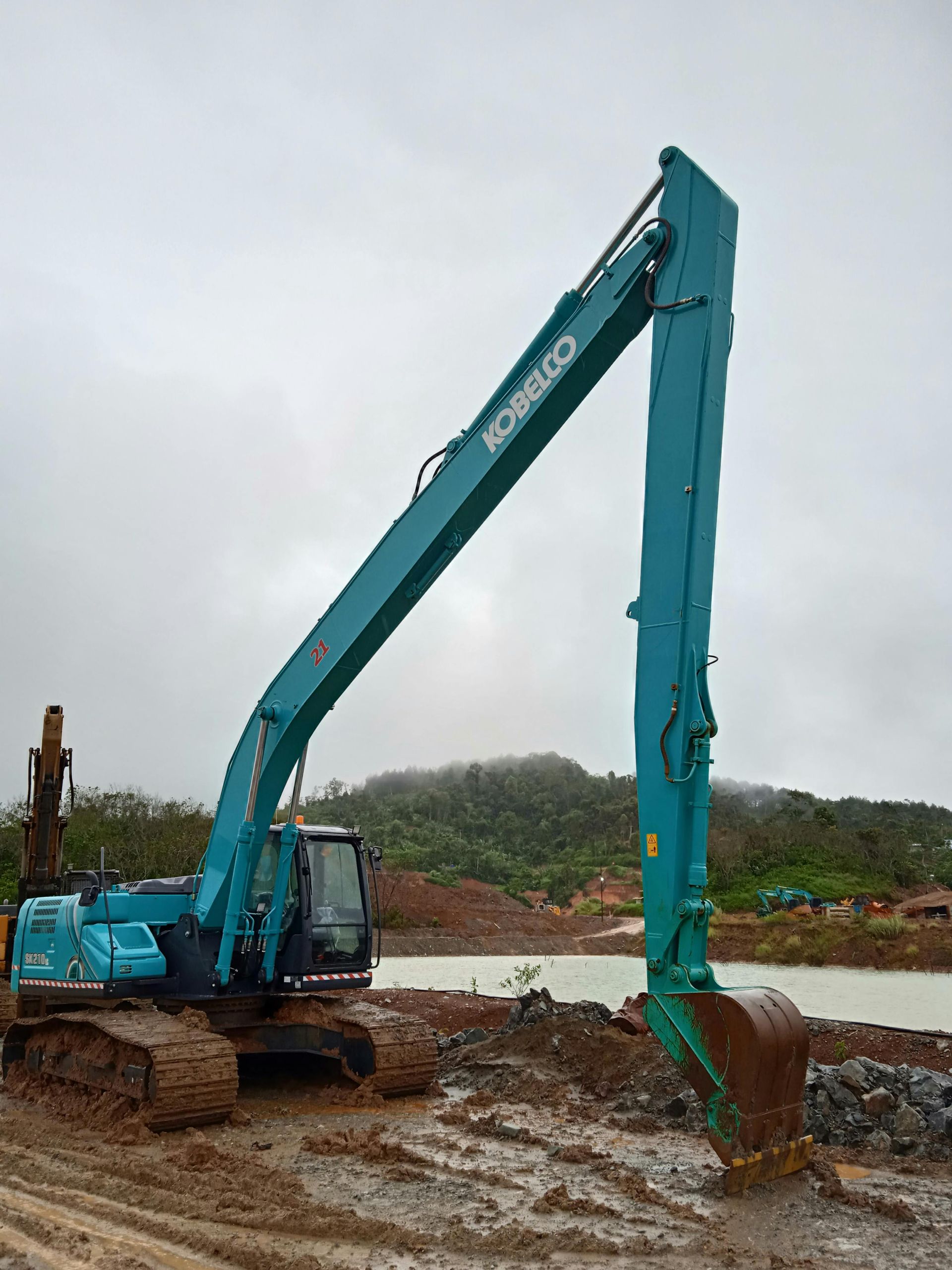 A teal Kobelco long-reach excavator sits on muddy ground at a construction site against a cloudy, mountainous background.