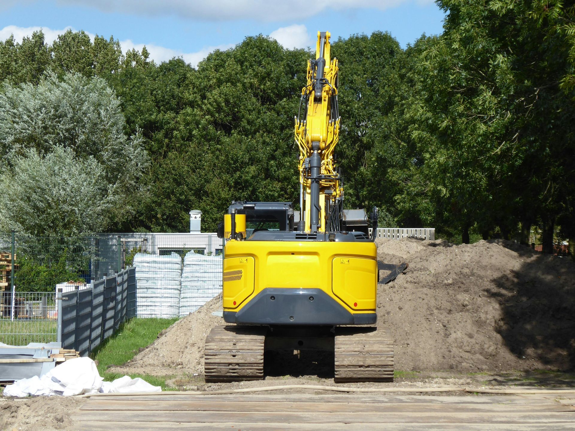 A bright yellow excavator sits on a dirt path in front of a pile of soil, with a line of trees in the background.