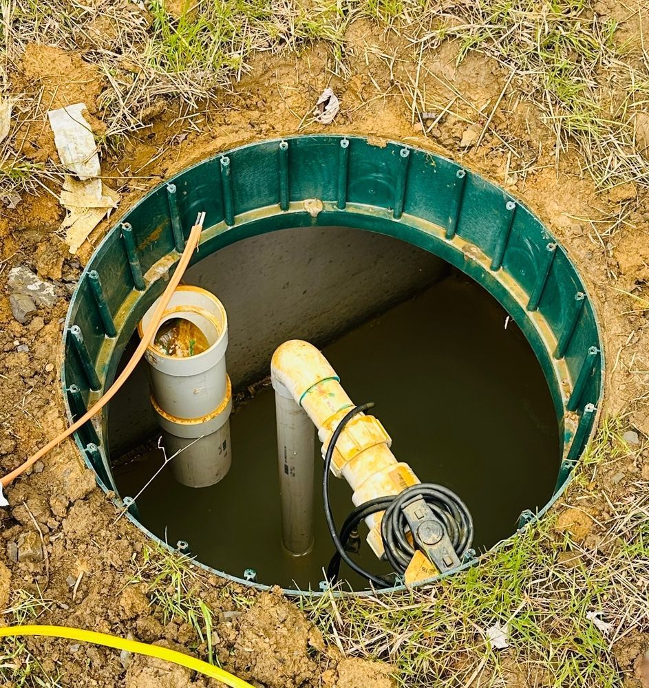 An open, green-rimmed underground utility or pump basin containing standing water, pipes, and electrical cables.