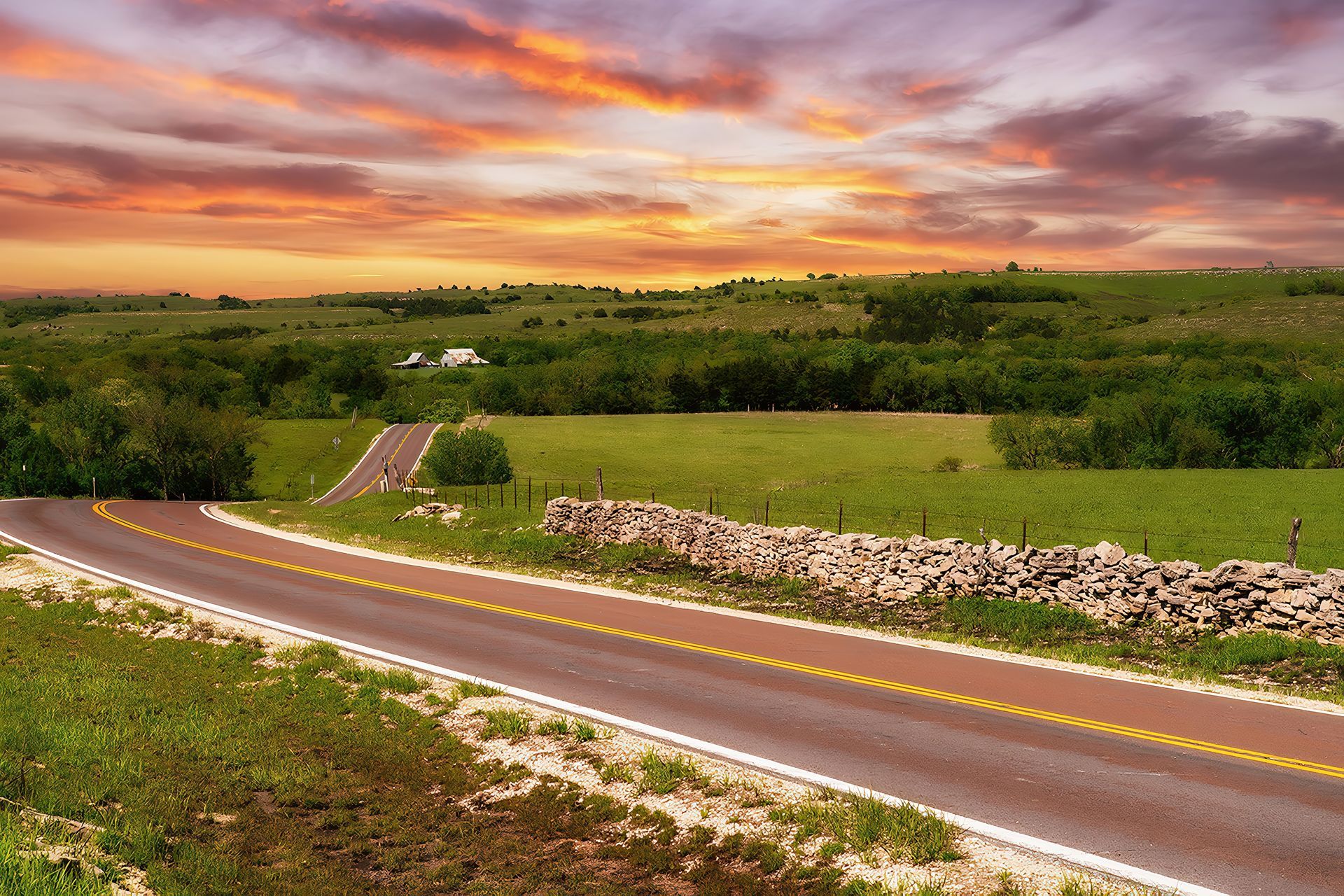 Winding country road through green fields under a dramatic orange sunset sky
