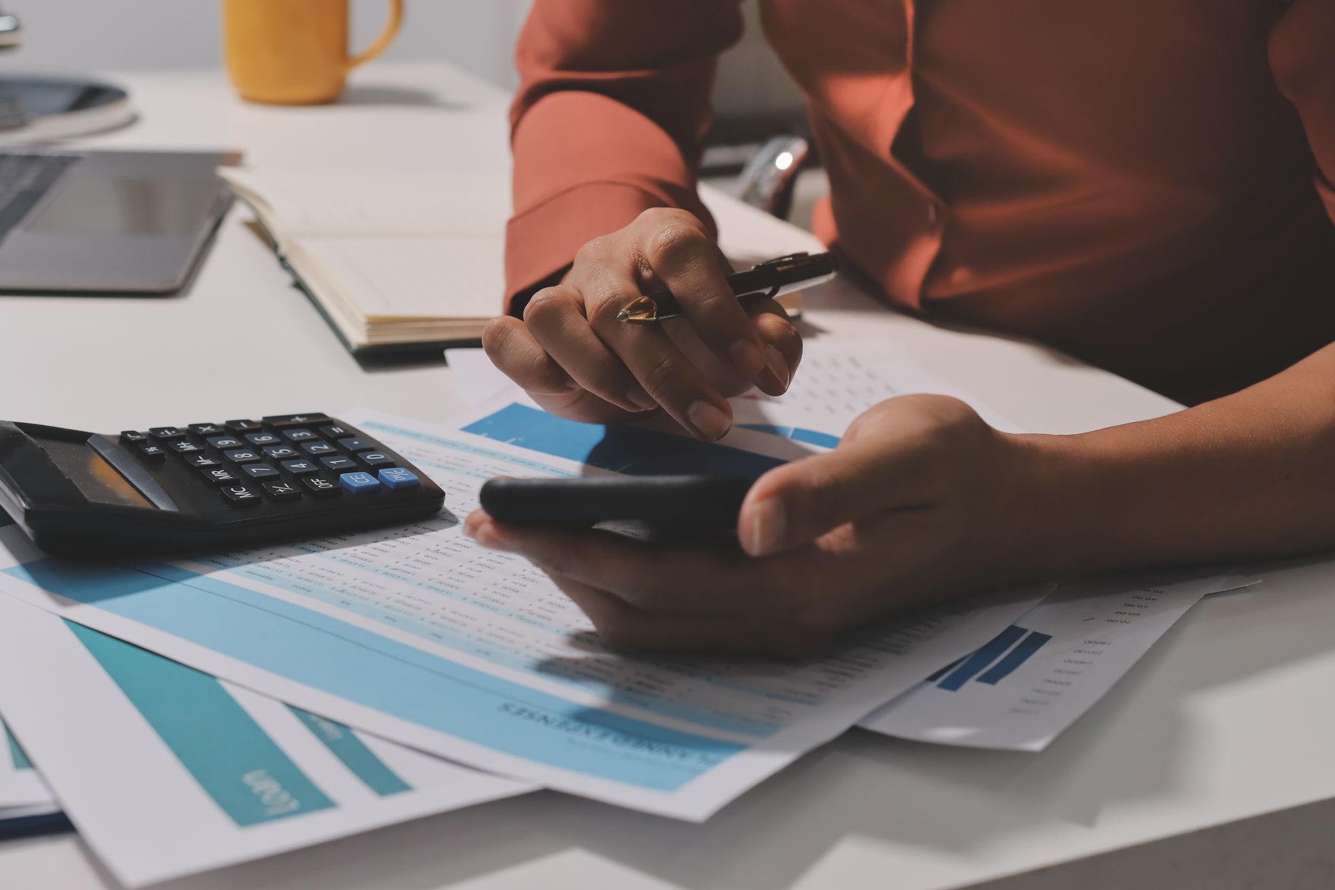 Person holding phone and pen, reviewing documents, calculator and mug on desk.